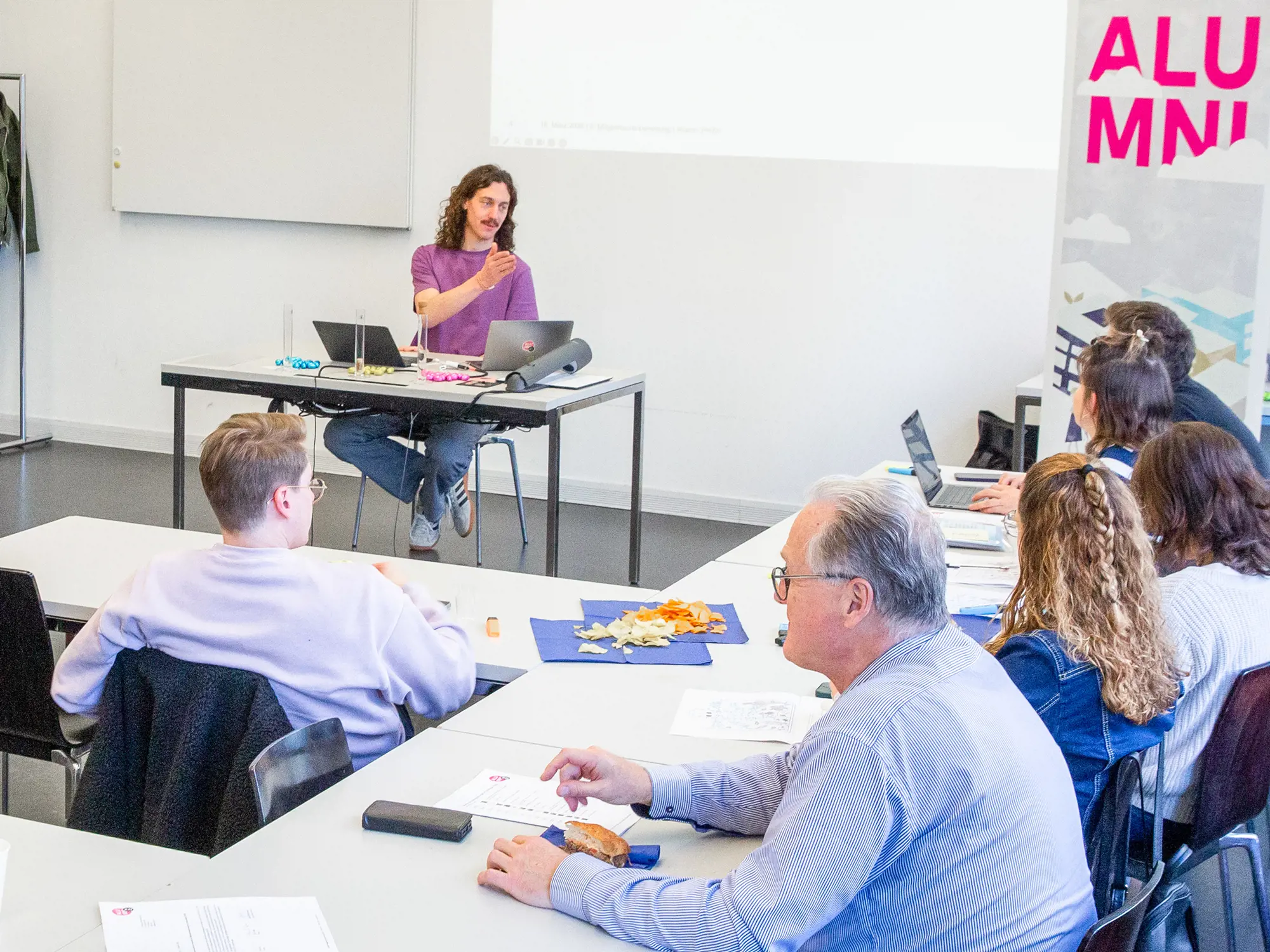 Ein Mann sitzt an einem Tisch mit Laptops und spricht zu einer Zuhörer:innengruppe in einem Seminarraum. Die Teilnehmenden sitzen an U-förmig angeordneten Tischen, hören aufmerksam zu und haben teilweise Snacks oder Unterlagen vor sich liegen. Am rechten Bildrand ist ein Roll-up-Banner mit der pinken Aufschrift «ALUMNI» zu sehen.