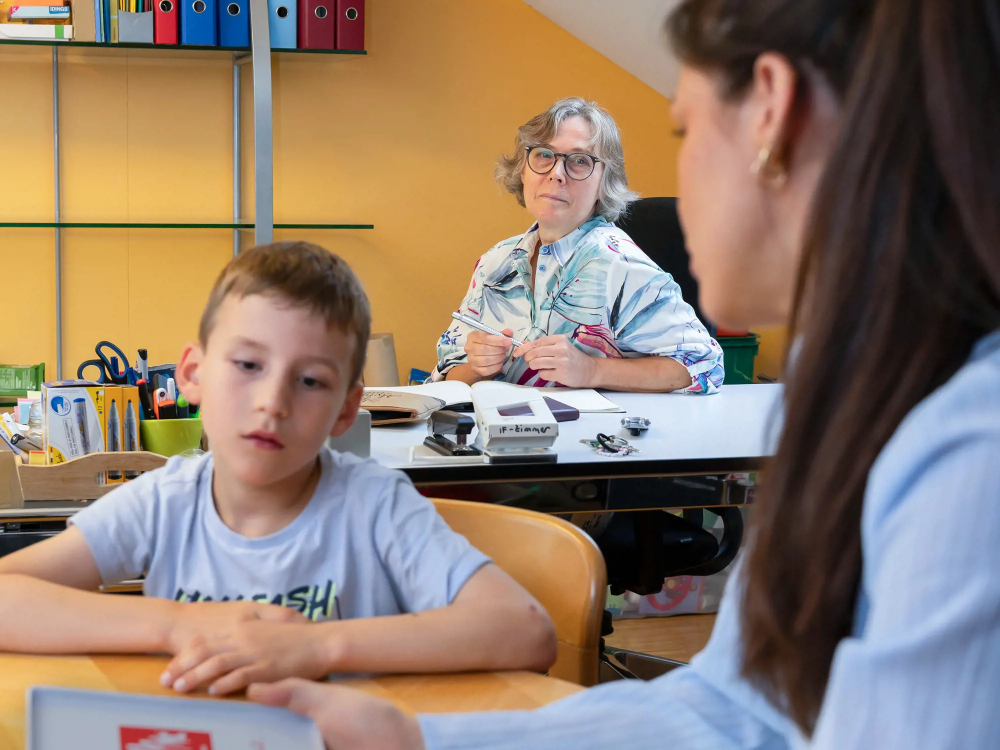 Das Bild zeigt eine Interaktionssituation in einem Klassenzimmer. Im Vordergrund sitzt ein Junge an einem Tisch und schaut ernsthaft zu einer jungen Frau, die mit dem Rücken zur Kamera sitzt und ein Buch hält. Im Hintergrund sitzt eine ältere Frau an einem Schreibtisch, schaut direkt in die Kamera und beobachtet die Szene.