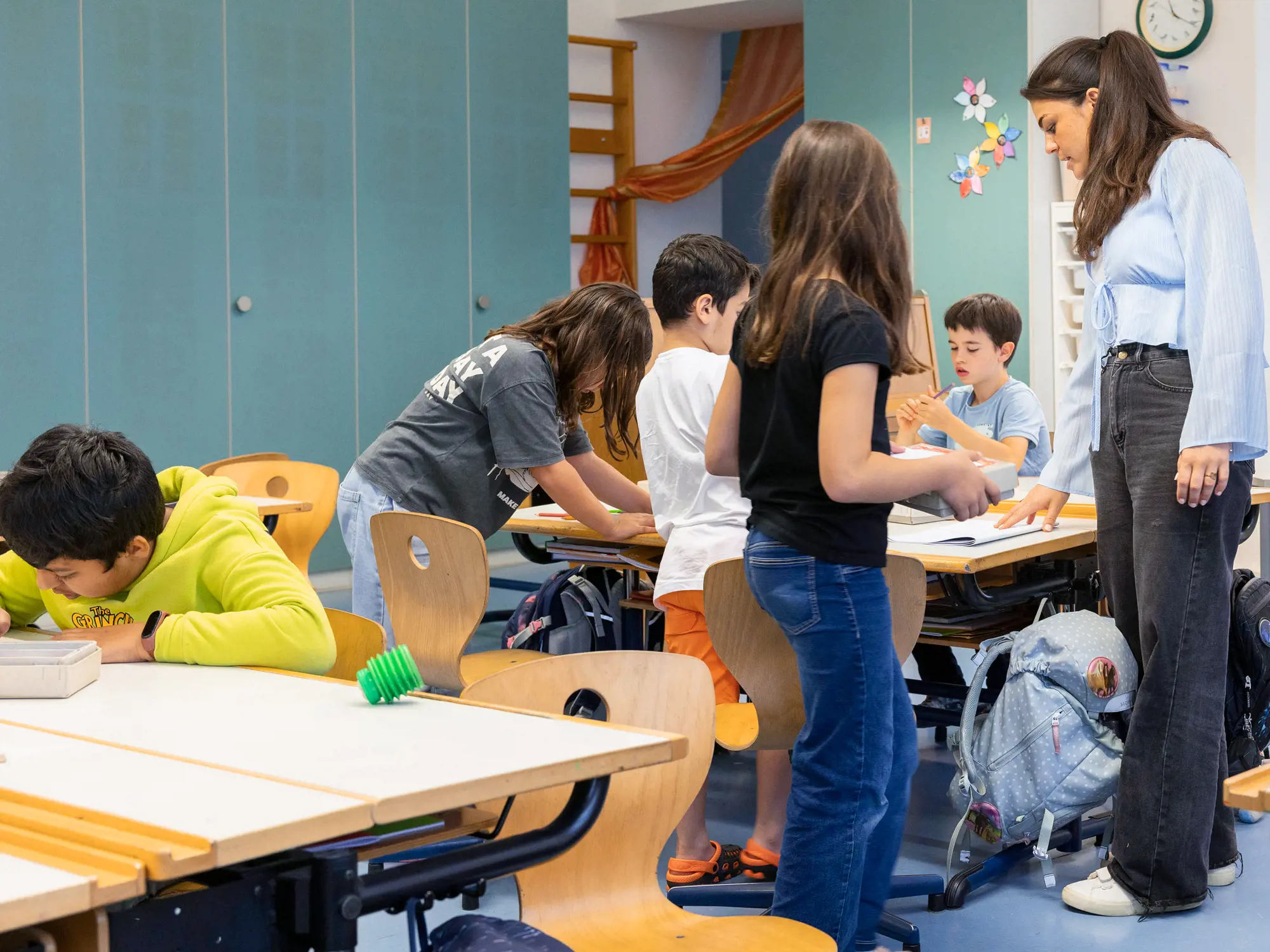 Das Bild zeigt eine lebhafte Szene in einem Klassenzimmer, in der mehrere Kinder in kleinen Gruppen an Schreibtischen arbeiten oder stehen. Eine junge Erwachsene steht bei einer Gruppe und beaufsichtigt oder leitet deren Aktivität. Die Kinder sind in verschiedene Aufgaben vertieft; einige beugen sich über den Tisch, andere schauen zu.
