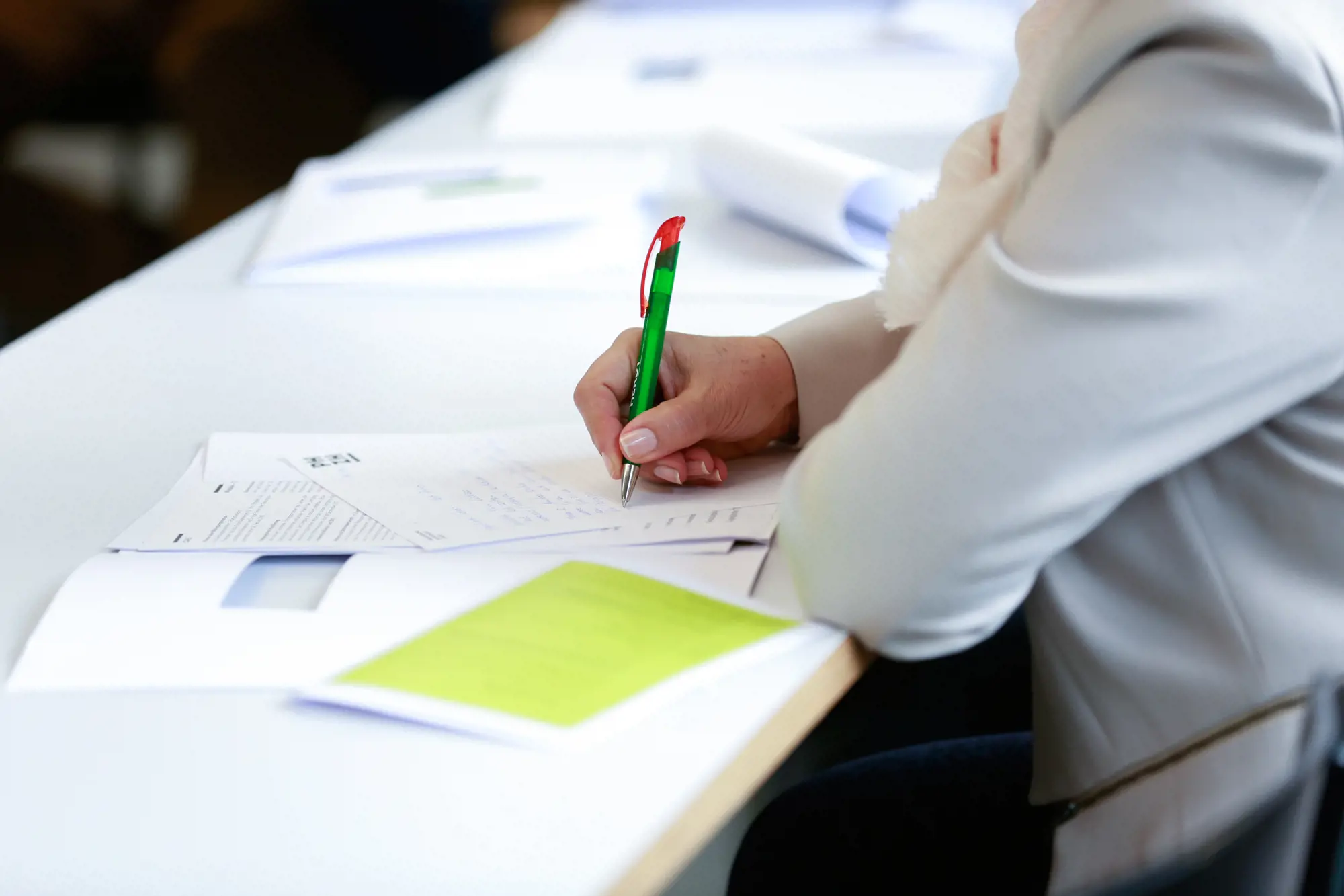 Eine Person sitzt an einem Tisch und schreibt mit einem grünen Kugelschreiber auf ein Papier. Der Kopf der Person ist nicht sichtbar.
