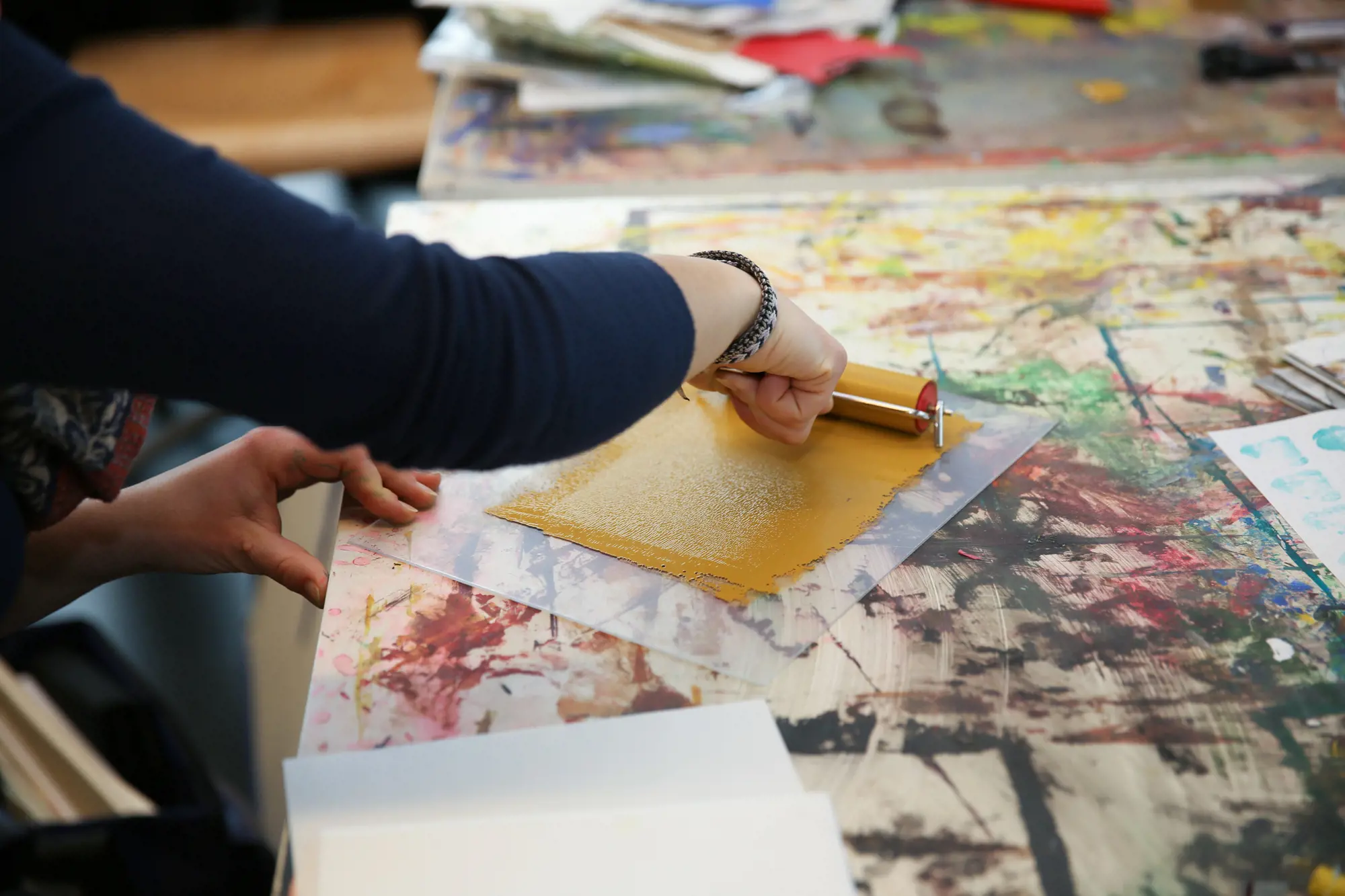 In a workshop, a person is rolling ochre-coloured paint onto a transparent surface on a work table covered in paint splatters.