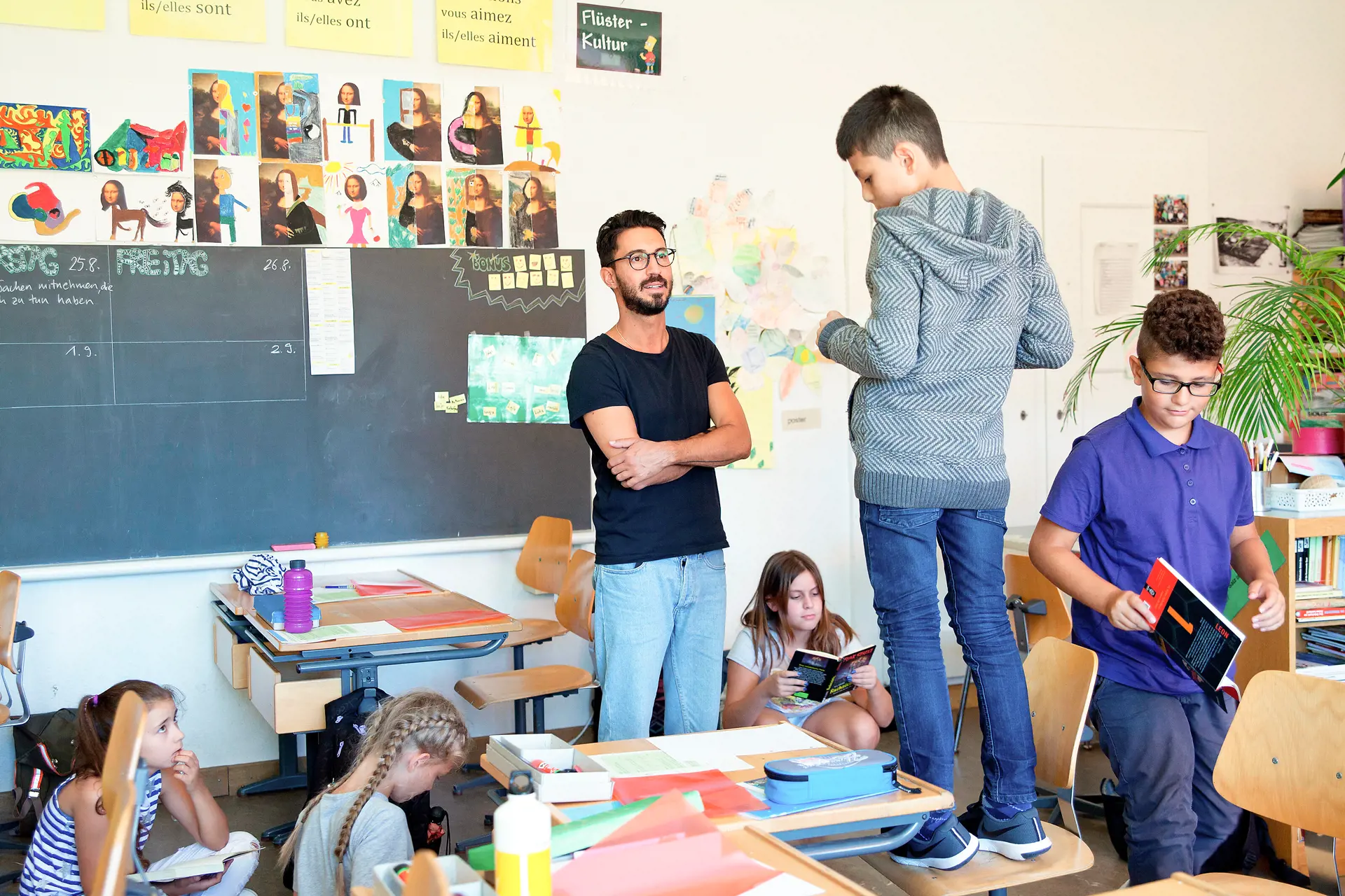 A school child stands on a chair in a classroom and talks to the classroom teacher.