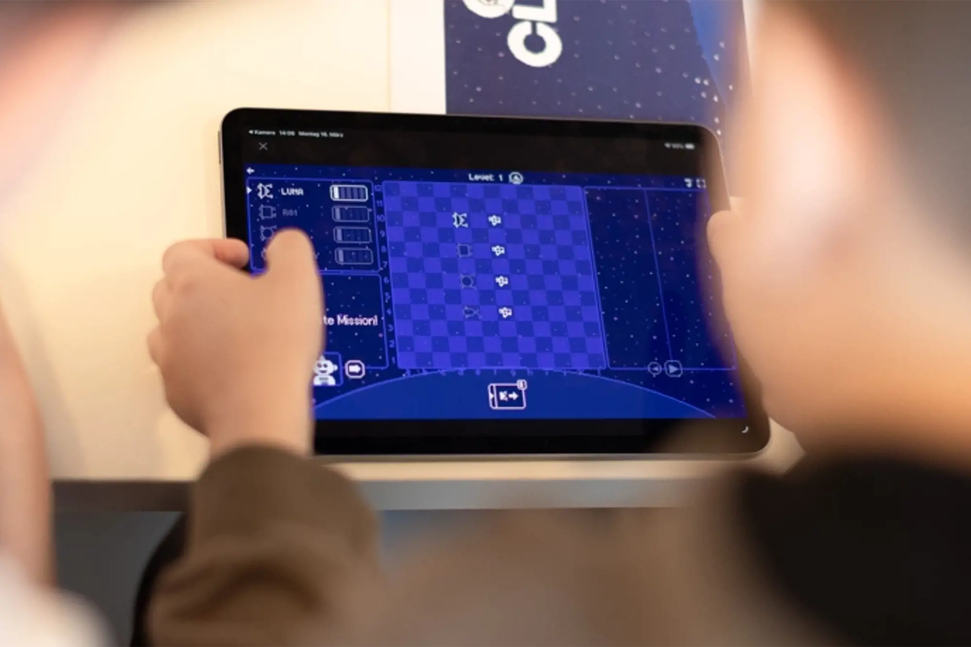 Eine Nahaufnahme über die Schulter einer jugendlichen Person zeigt ein Tablet, das mit beiden Händen gehalten wird. Auf dem Bildschirm ist eine blaue, weltraumbezogene Spieloberfläche mit einem schachbrettartigen Spielfeld, Symbolen und Bedienelementen zu sehen.