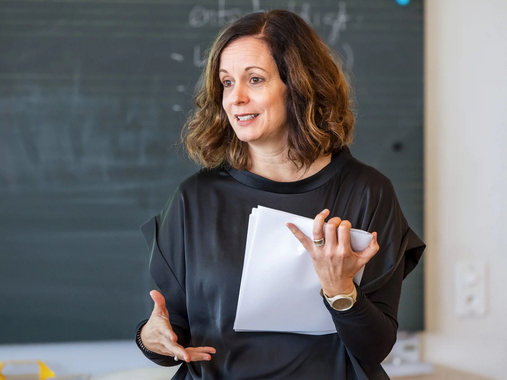 Das Foto zeigt eine Frau mit schulterlangen, brünetten Haaren und einem schwarzen Oberteil, die in einem hellen Raum vor einer dunklen Wandtafel steht. Sie hält weisse Papierblätter in der linken Hand und gestikuliert mit der rechten, während sie spricht.