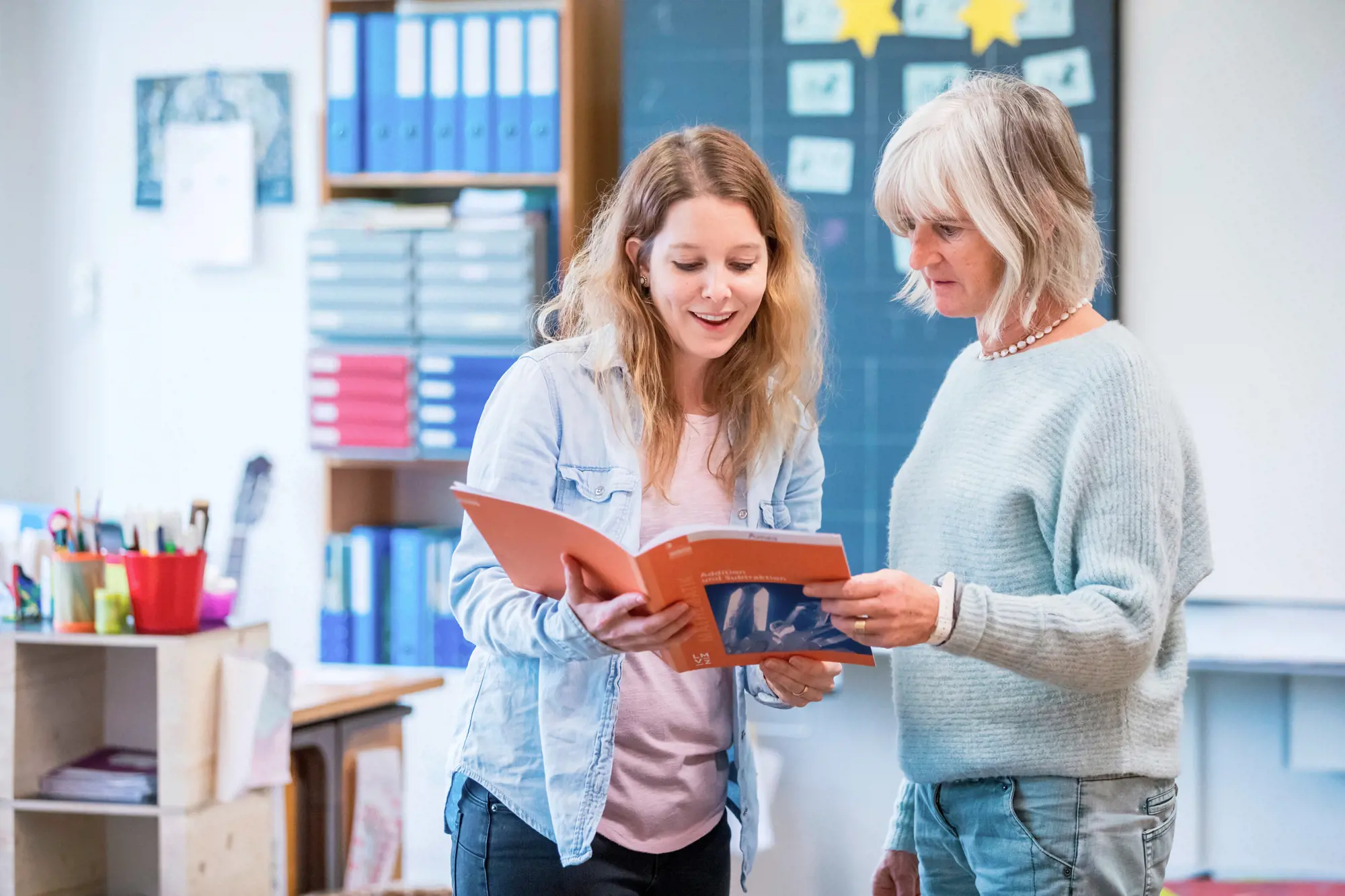 Eine ältere und eine jüngere Lehrerin blicken stehend in ein Lehrmittel.
