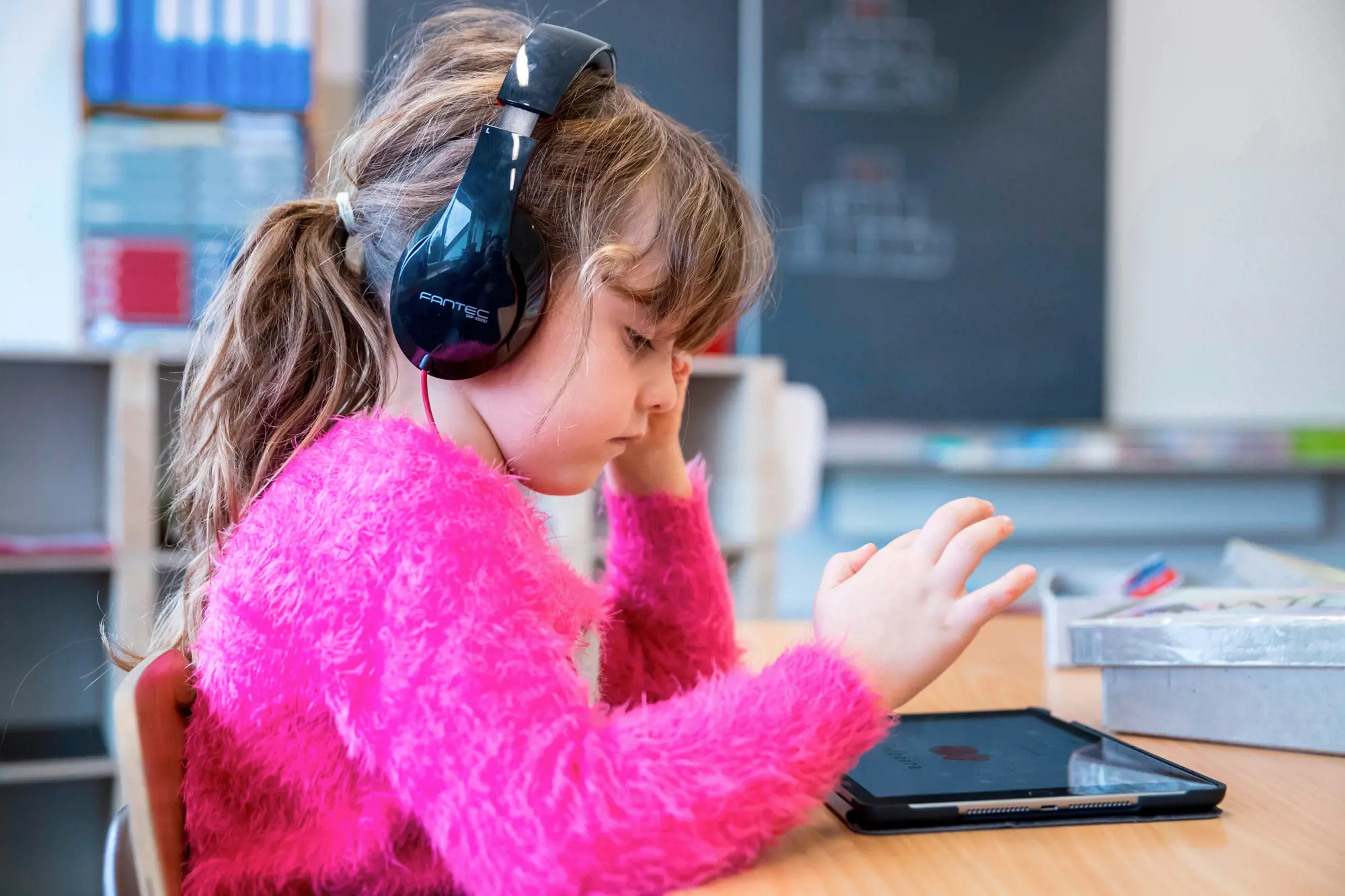 A girl with headphones and a pink jumper is sitting in front of a tablet.