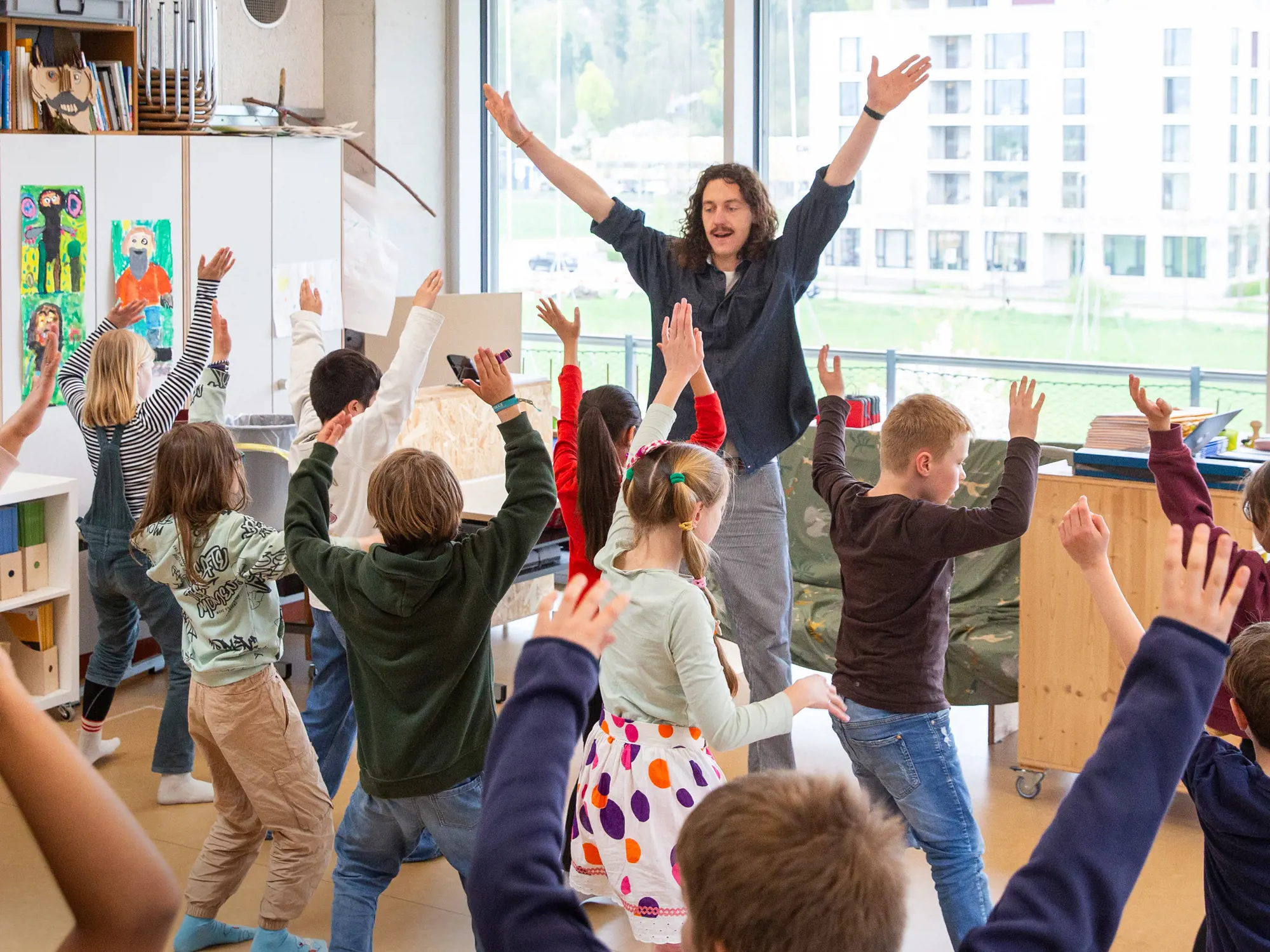 Ein Mann steht mit weit nach oben ausgestreckten Armen vor einer Gruppe von Kindern in einem hellen Klassenzimmer. Die Kinder stehen im Kreis um ihn herum und machen die Bewegung mit erhobenen Armen begeistert nach. Im Hintergrund gewährt eine Fensterfront den Blick auf eine Wiese und ein Gebäude im Freien.