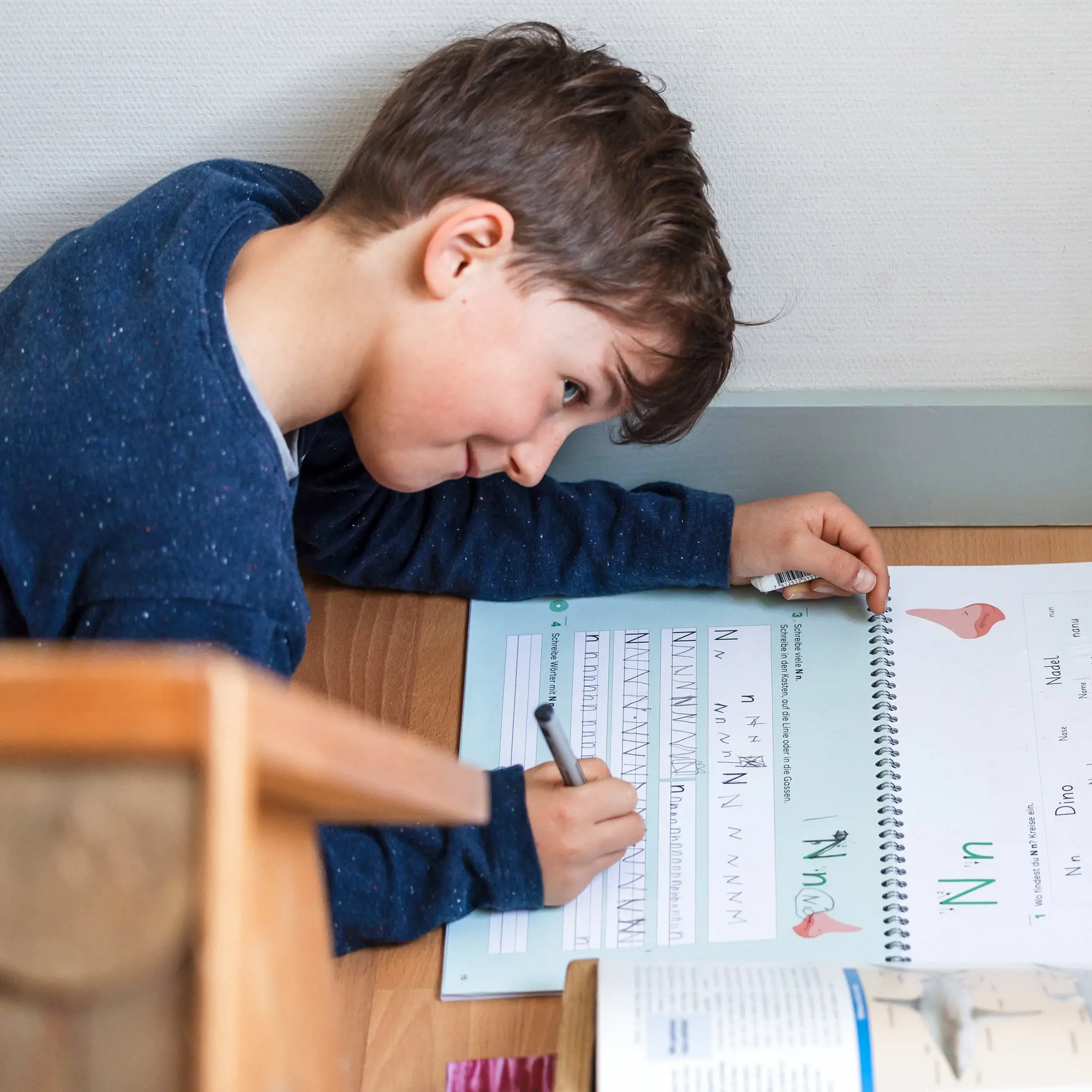 Ein auf dem Boden liegendes Kind beugt sich über ein Übungsheft und schreibt konzentriert mit einem Stift. Auf den Buchseiten sind Schreibübungen für den Buchstaben «N» sowie Zeichnungen und Wörter wie «Nadel» oder «Dino» zu sehen.
