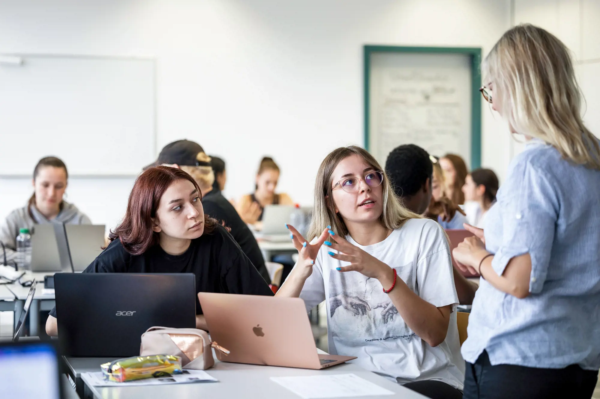 Zwei Berufsschülerinnen sitzen an einem Tisch und hören aufmerksam ihrer Lehrerin zu, die vor ihnen steht und auf dem Bild nur von hinten sichtbar ist.