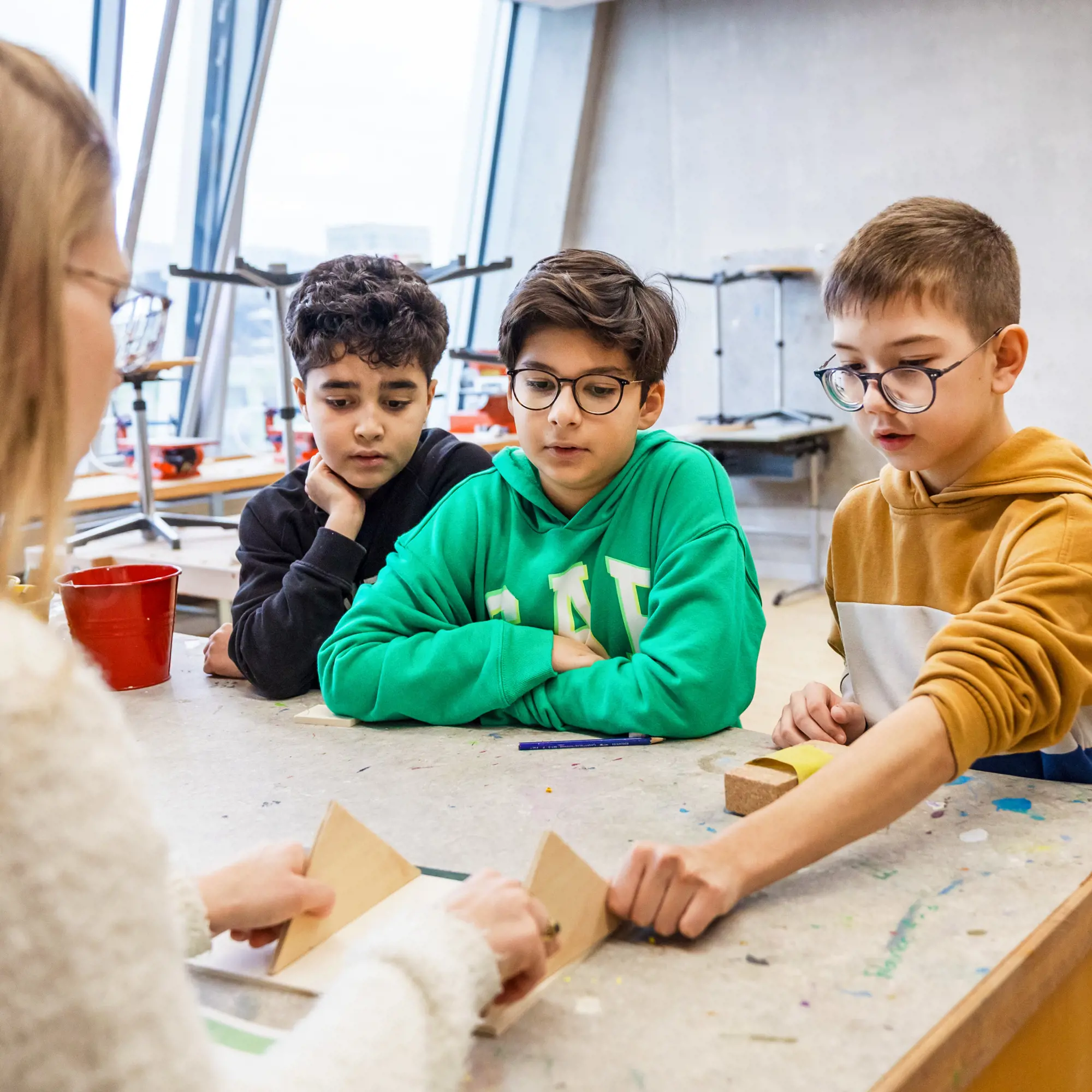 Das Foto zeigt drei Jungen, die an einem Arbeitstisch in einem Werkraum sitzen und konzentriert eine Lehrerin bei der Arbeit mit Holzstücken beobachten. Sie hält zwei Holzdreiecke auf der Tischplatte zusammen. Die drei Schüler verfolgen den Vorgang aufmerksam: Ein Junge in einem grünen Kapuzenpullover sitzt in der Mitte, links daneben ein Junge in Schwarz, der seinen Kopf auf die Hand stützt, und rechts ein Junge in einem senfgelben Hoodie, der mit seiner Hand ebenfalls nach einem Materialstück auf dem Tisch greift. Die Tischoberfläche weist bunte Farbflecken auf, und im Hintergrund sind durch grosse Fensterfronten weitere Werkbänke erkennbar.