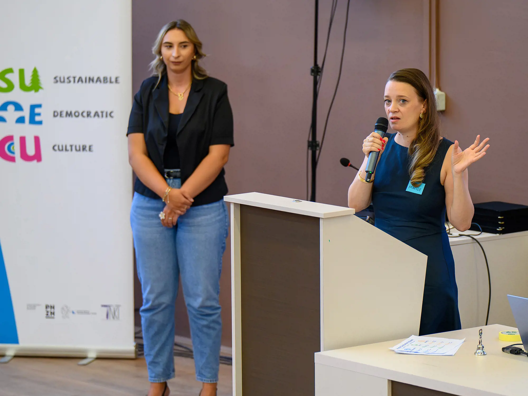 Magdalena Spasovska spricht an einem Rednerpult mit einem Mikrofon; neben ihr steht eine weitere Frau vor einem SUDECU-Banner.
