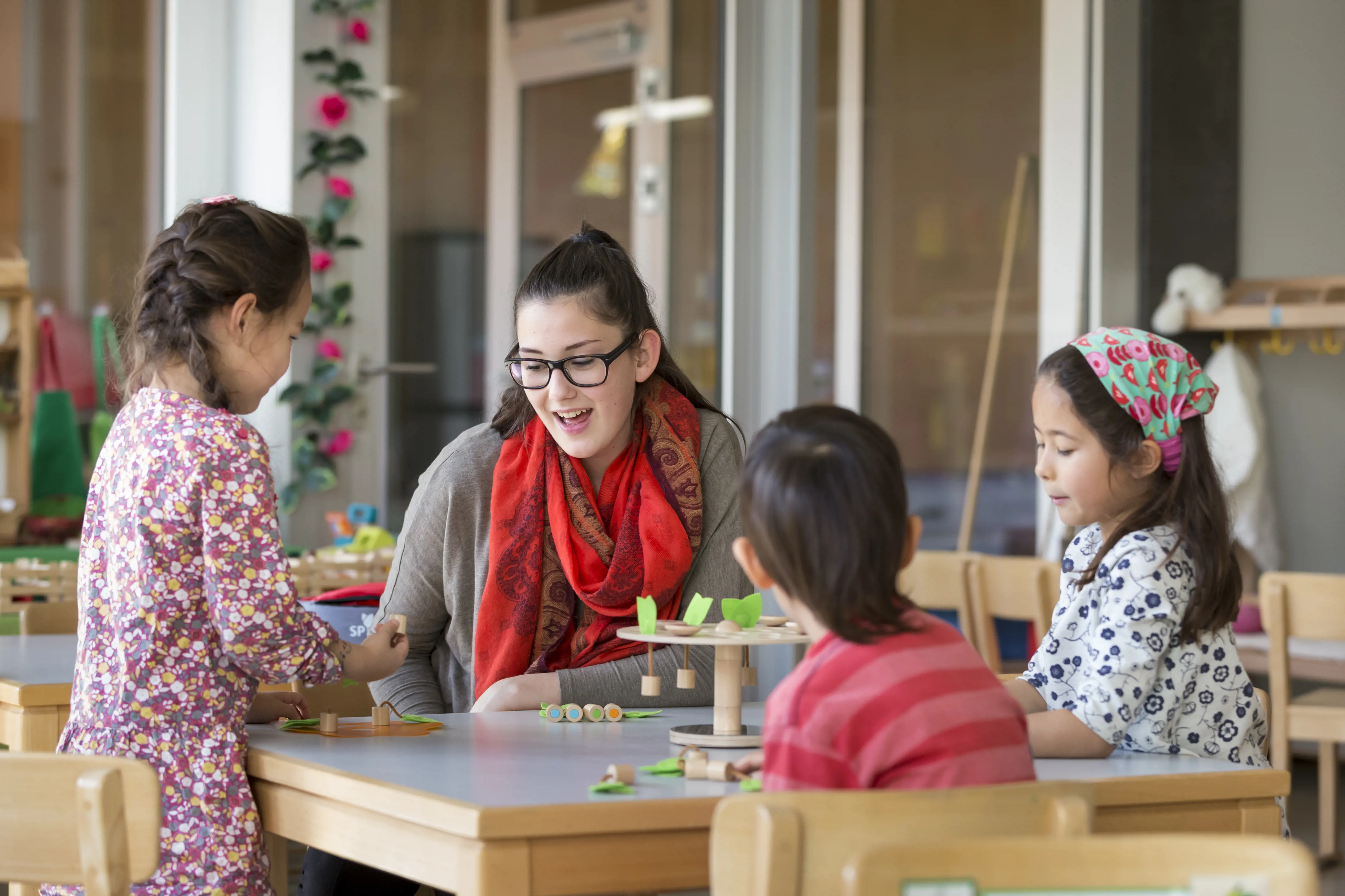 Eine Lehrerin sitzt am Tisch mit drei Kindern, die am basteln sind.