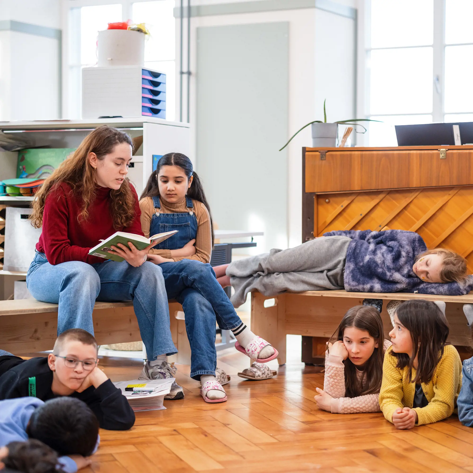 Eine Lehrerin sitzt auf einer Holzbank und liest einer Gruppe von Kindern aus einem Buch vor. Einige Kinder liegen oder sitzen aufmerksam auf dem Parkettboden, während ein anderes Kind entspannt auf einer Bank im Hintergrund liegt. Die gesamte Atmosphäre wirkt ruhig, konzentriert und gemütlich.
