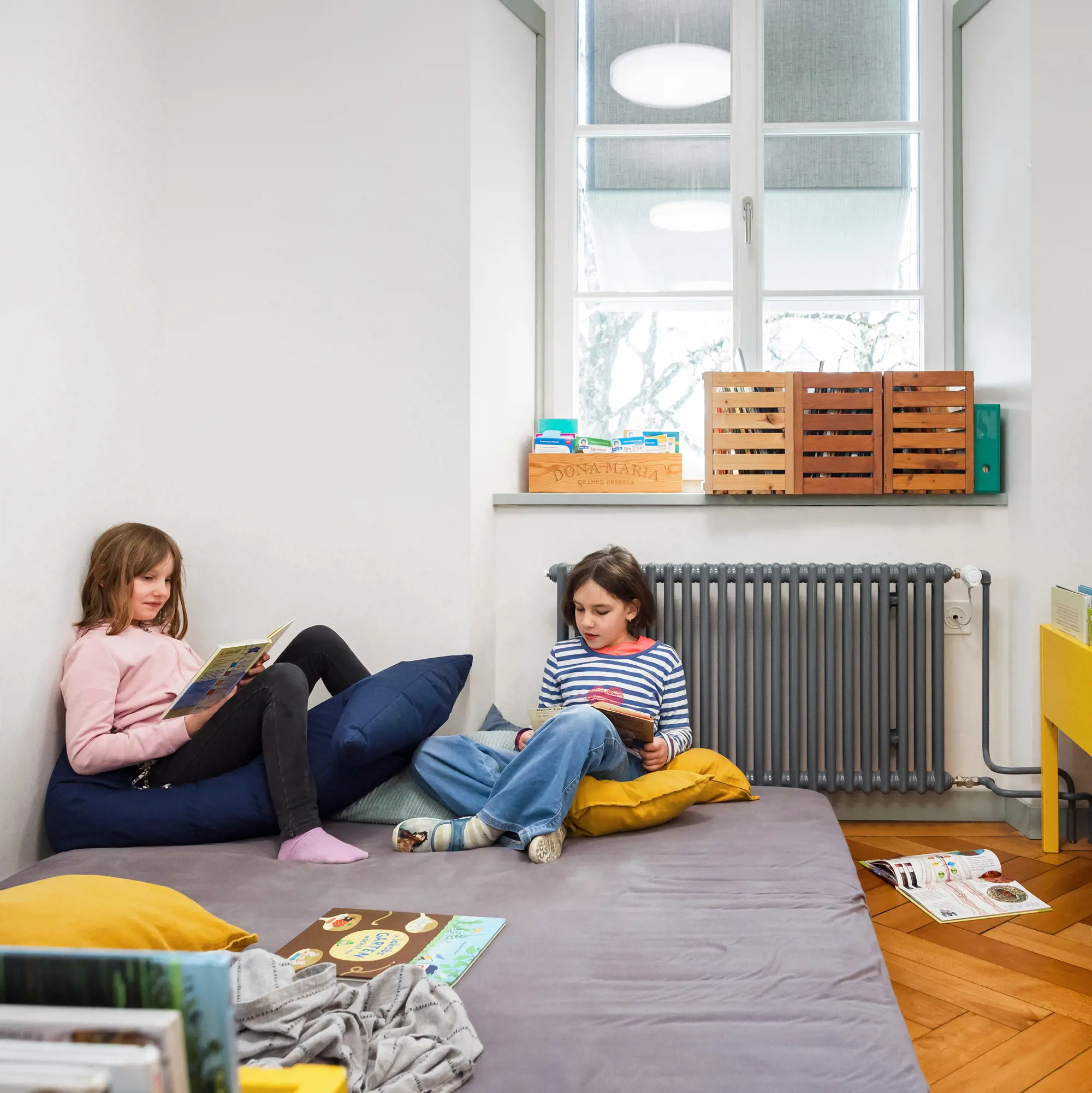 Zwei Kinder sitzen gemütlich auf einer grossen, grauen Matte am Boden in einer Leseecke und lesen konzentriert in ihren Büchern. Sie sind von Kissen umgeben, während im Raum ein Fenster mit Holzboxen auf dem Sims, ein Heizkörper und weitere Bücher auf dem Parkettboden zu sehen sind. Die Atmosphäre wirkt ruhig und einladend.