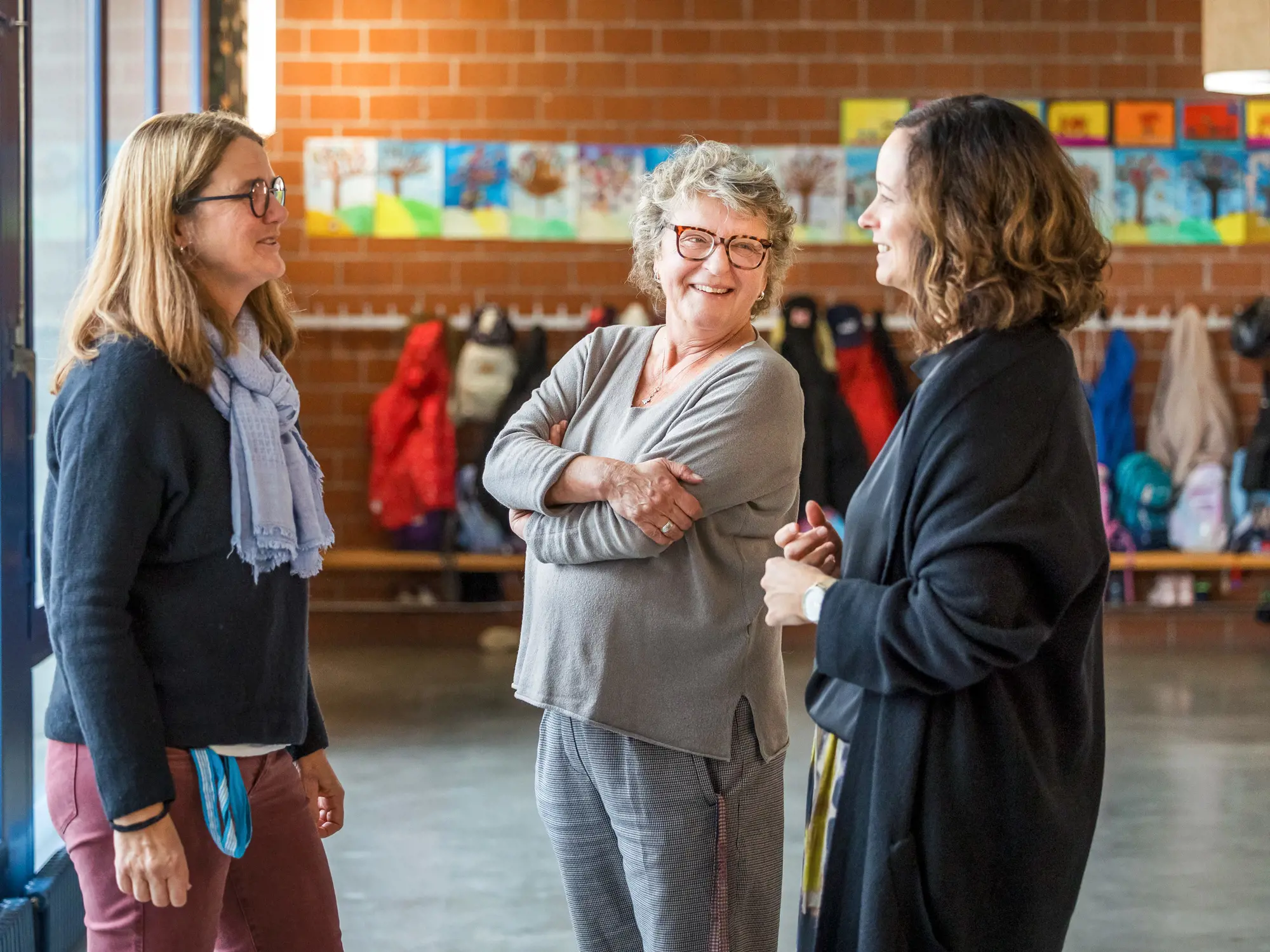 Das Foto zeigt drei Frauen, die in einem entspannten und freundlichen Gespräch in einem Schulflur zusammenstehen. Die Frau in der Mitte trägt ein graues Oberteil und eine Brille; sie lächelt herzlich mit verschränkten Armen. Die beiden anderen Frauen stehen ihr gegenüber und beteiligen sich aktiv am Dialog. Im Hintergrund ist eine typische Garderobe mit Kinderjacken vor einer roten Backsteinwand zu erkennen, an der bunte Kinderzeichnungen hängen.