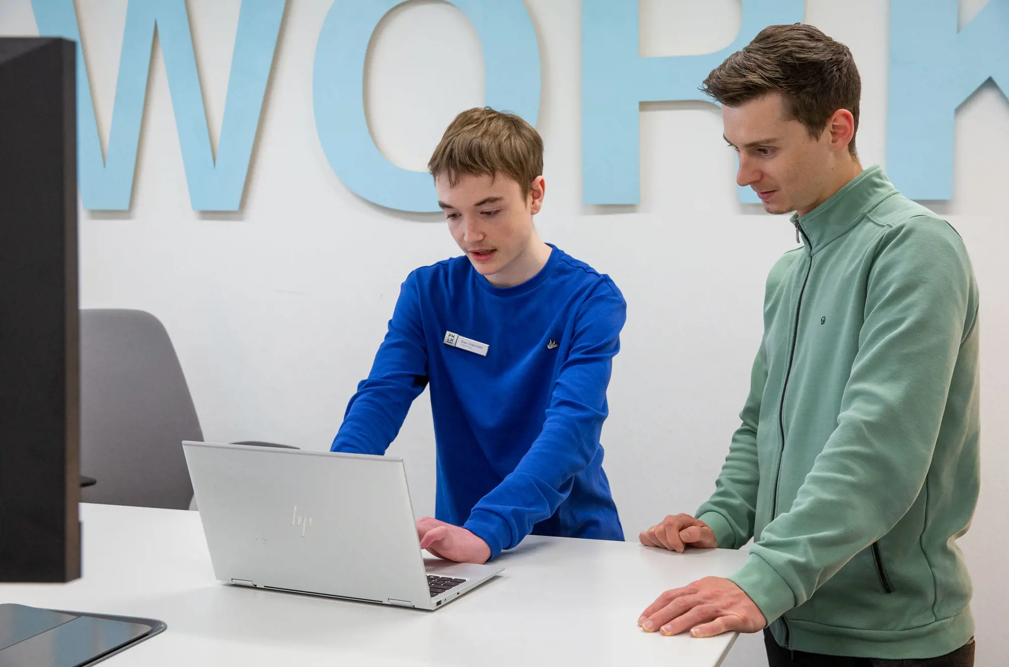 Ein Lernender ICT-Fachmann steht mit einem PHZH-Mitarbeiter vor seinem Laptop und erklärt ihm etwas.