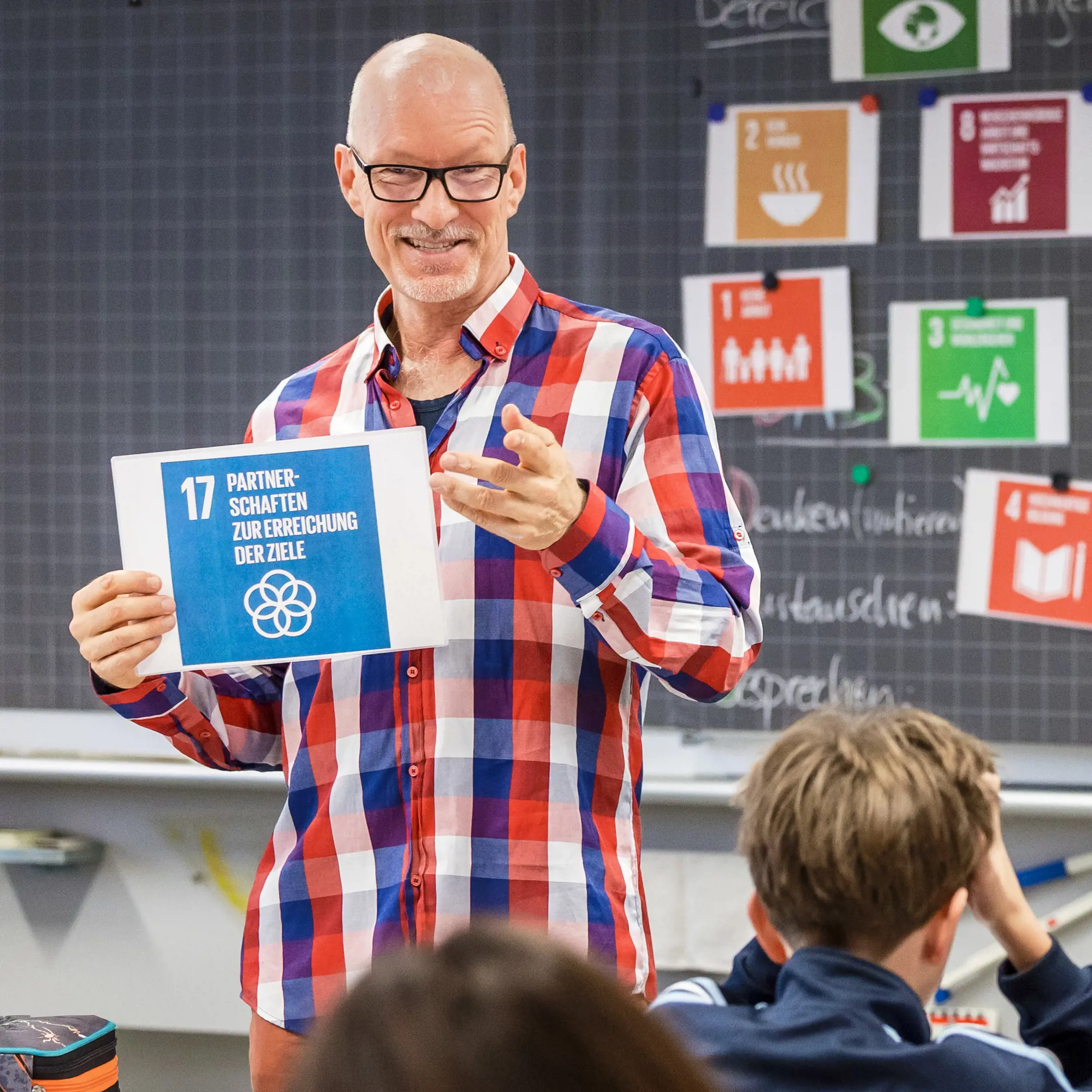 Ein lächelnder Lehrer mit Brille und kariertem Hemd steht vor einer Wandtafel und hält eine Infokarte hoch. Auf der blauen Karte steht «17 Partnerschaften zur Erreichung der Ziele». Im Hintergrund hängen weitere bunte Symbole der UN-Nachhaltigkeitsziele an der Tafel, während im Vordergrund die Köpfe von Schülern im Unterricht zu sehen sind.