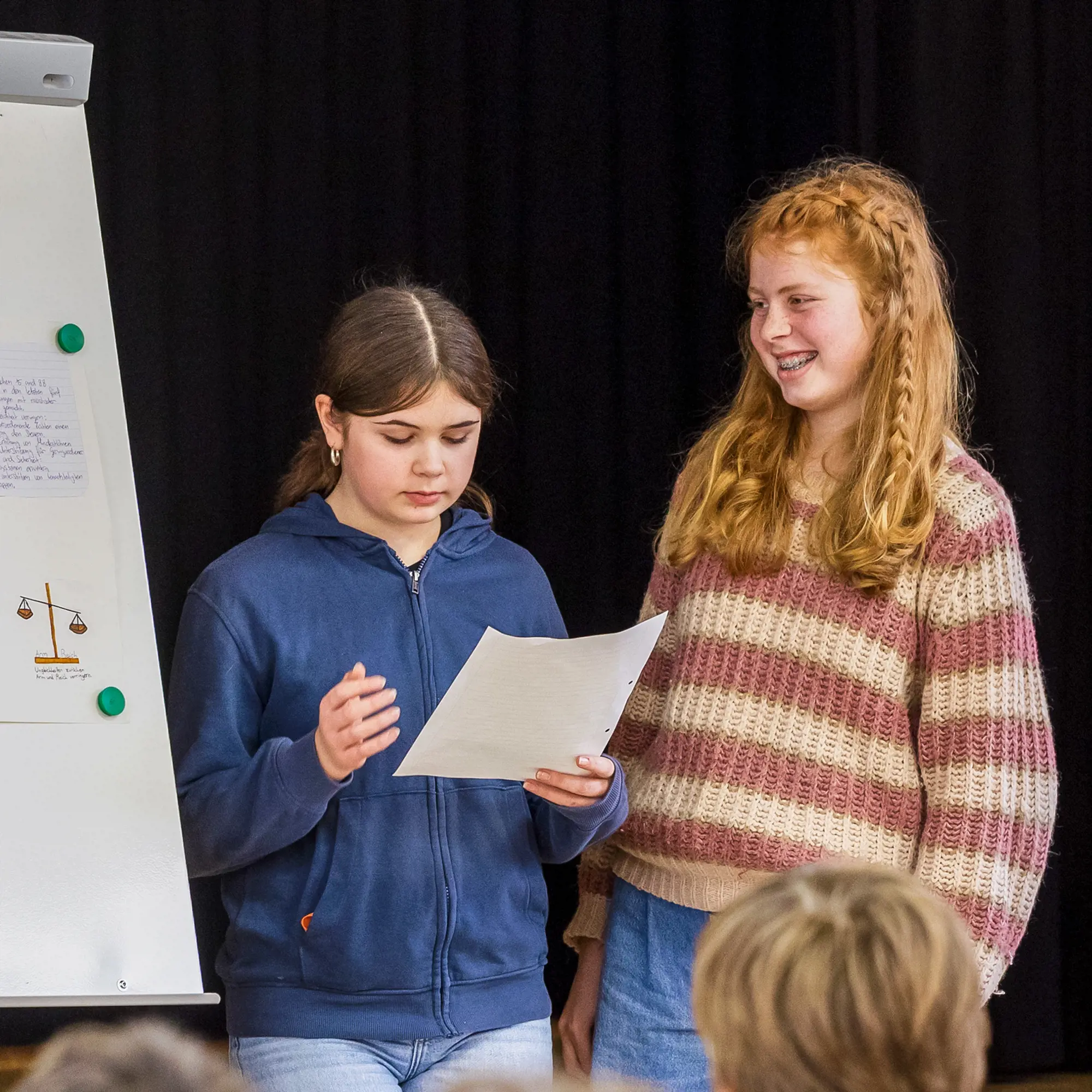 Zwei Schülerinnen stehen vor einem Flipchart und präsentieren der Klasse ihre Ergebnisse. Eine Schülerin liest konzentriert von einem Blatt Papier ab, während die andere sie lächelnd ansieht. Auf dem Flipchart ist eine Zeichnung einer Waage zu erkennen.