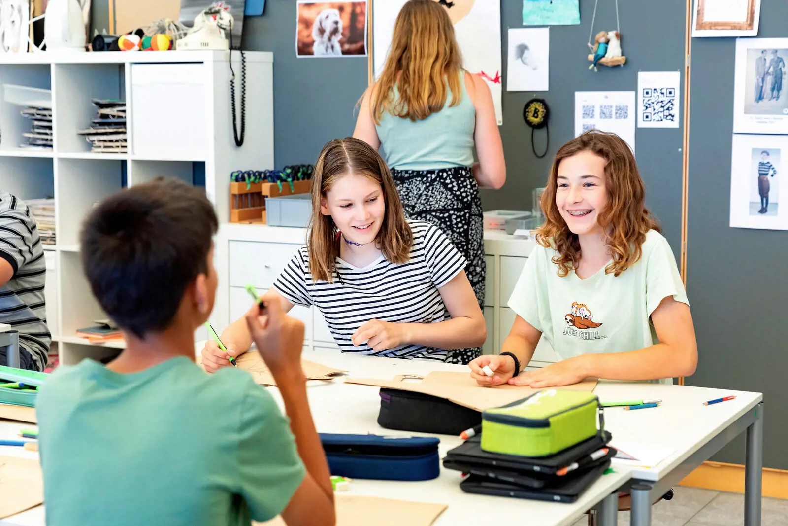Zwei Schülerinnen (von vorne sichtbar) und ein Schüler (von hinten sichtbar) sitzen um einen Tisch.