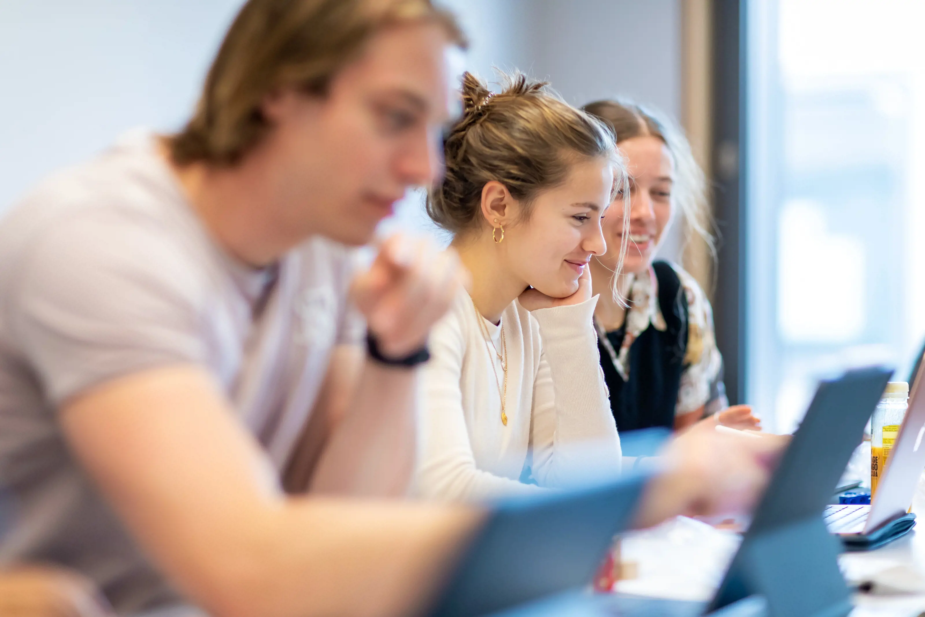 Ein Student und zwei Studentinnen sitzen an ihren Laptops während eines Moduls. Die mittlere Studentin lächelt.