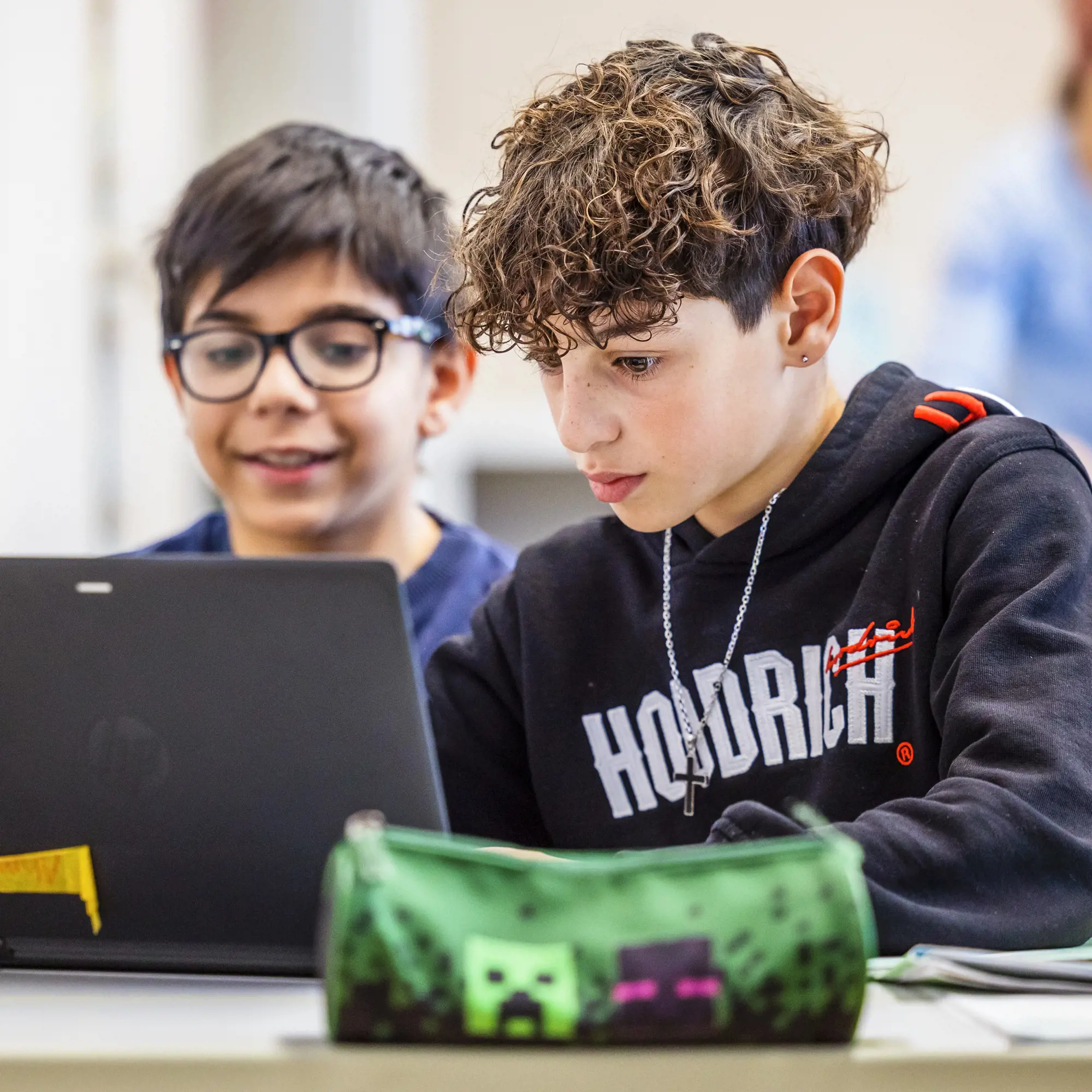 Das Foto zeigt zwei Jugendliche in einem Klassenzimmer, die gemeinsam auf einen Laptop-Bildschirm blicken. Im Vordergrund sitzt ein Junge mit lockigem braunem Haar und einem schwarzen Kapuzenpullover. Er blickt konzentriert auf den Computer. Direkt hinter ihm ist ein zweiter Junge mit Brille zu sehen, der lächelnd ebenfalls das Geschehen auf dem Bildschirm verfolgt.