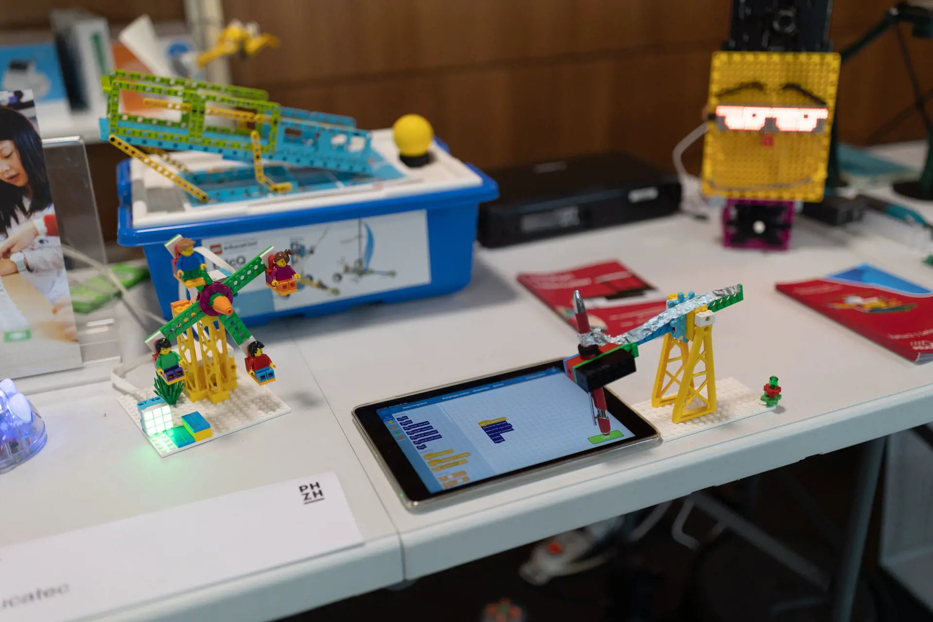 Auf einem Arbeitstisch sind verschiedene farbenfrohe Modelle aus Klemmbausteinen und Robotik-Elementen aufgebaut. Im Vordergrund liegt ein Tablet mit einer Programmieroberfläche, das mit einer kleinen Kran-Konstruktion aus Klemmbausteinen verbunden ist. Links daneben steht ein buntes Miniatur-Riesenrad mit kleinen Spielfiguren, während im Hintergrund weitere Lern- und Baukästen sowie eine gelbe Figur mit LED-Anzeige zu sehen sind.