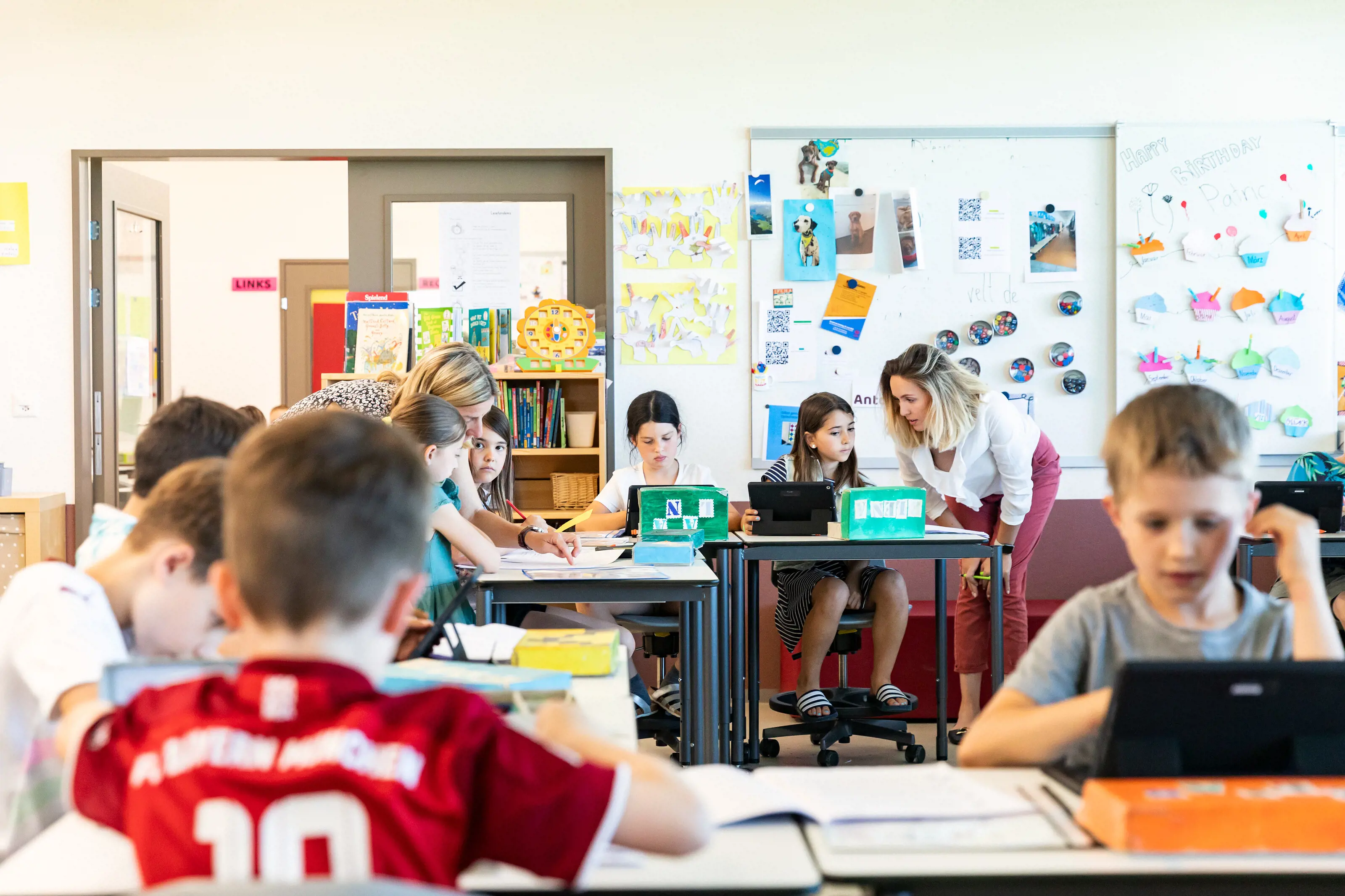 Schülerinnen und Schüler sitzen an ihren Tischen. Zwei Lehrer:innen beugen sich zu den Schüler:innen und sprechen mit ihnen.