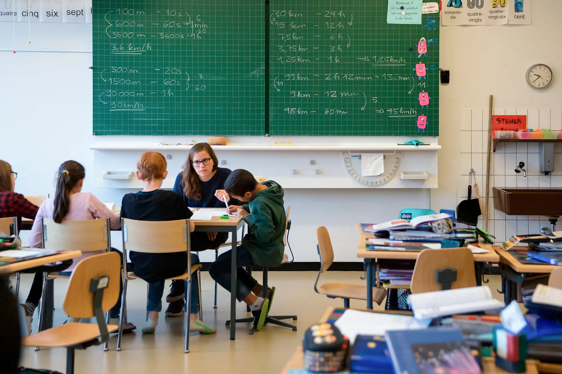Four children are sitting with a teacher at a table in front of a blackboard. Mathematics tasks can be seen on it.