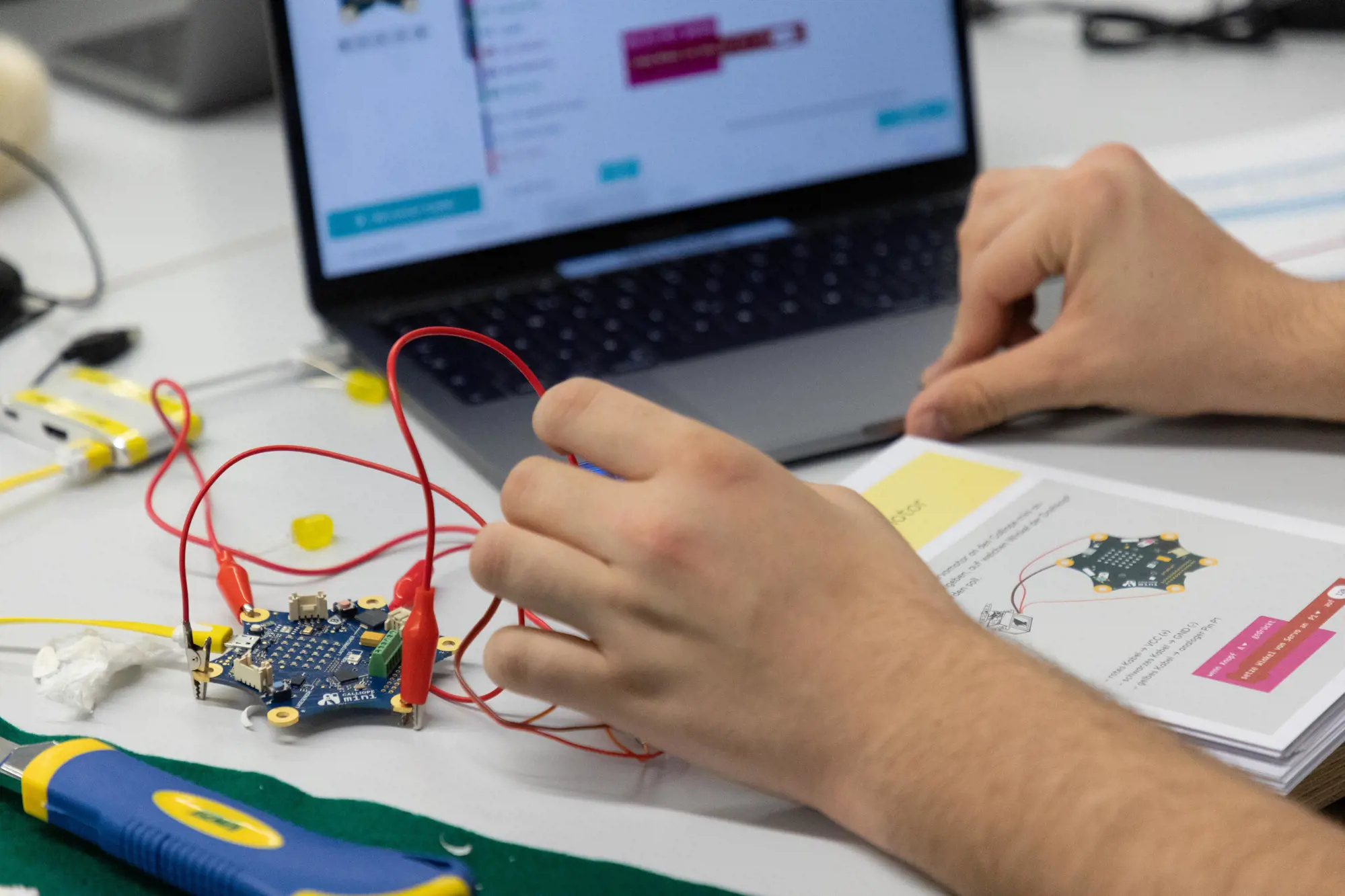 On a table there is a Calliope mini, a manual for it and a laptop, and in front of it are the hands of a person.
