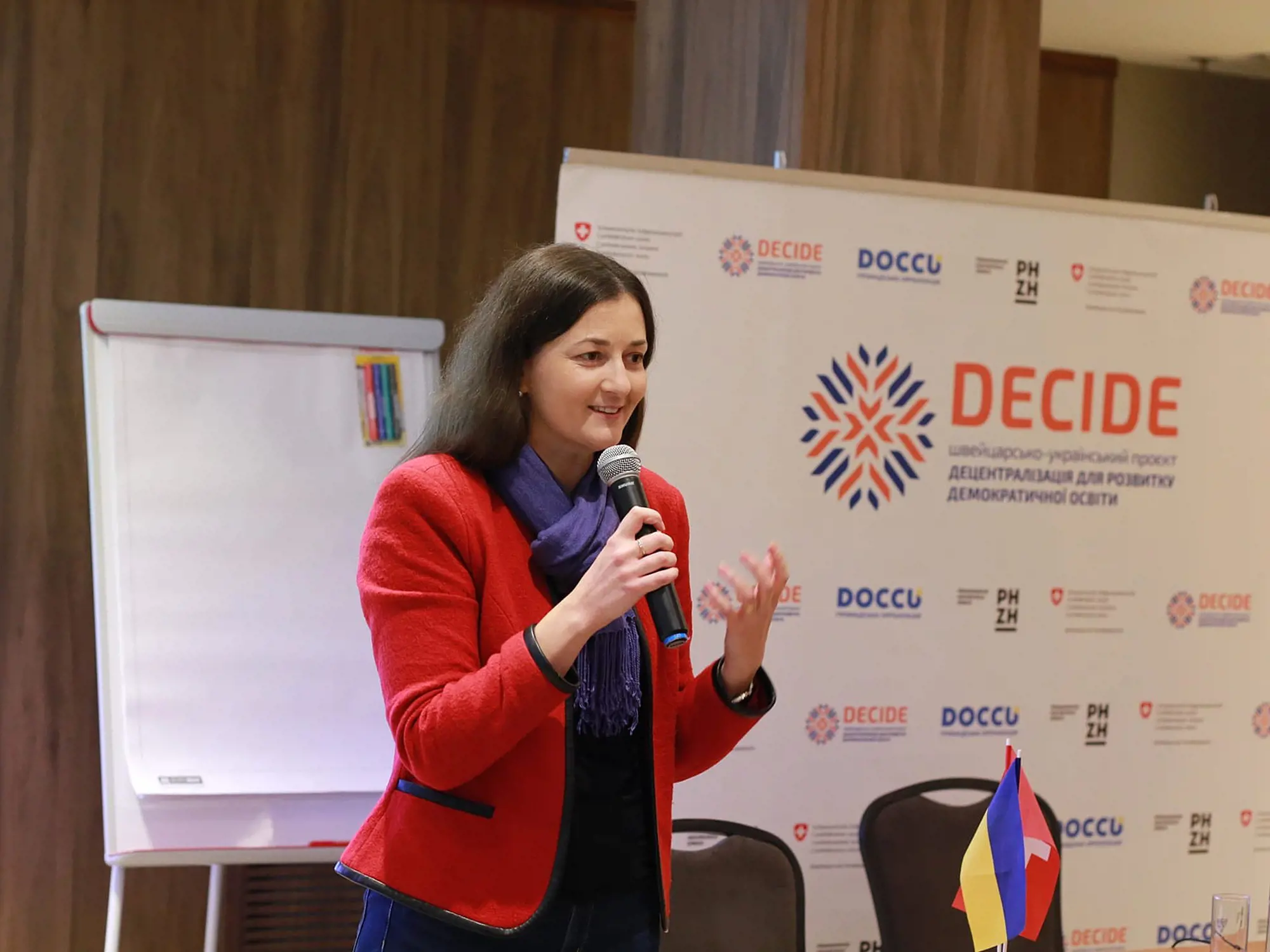 llona Postemska, Senior National Programme Officer der Schweizer Botschaft in der Ukraine in einem roten Jackett hält ein Mikrofon in der Hand und spricht vor einem Banner.