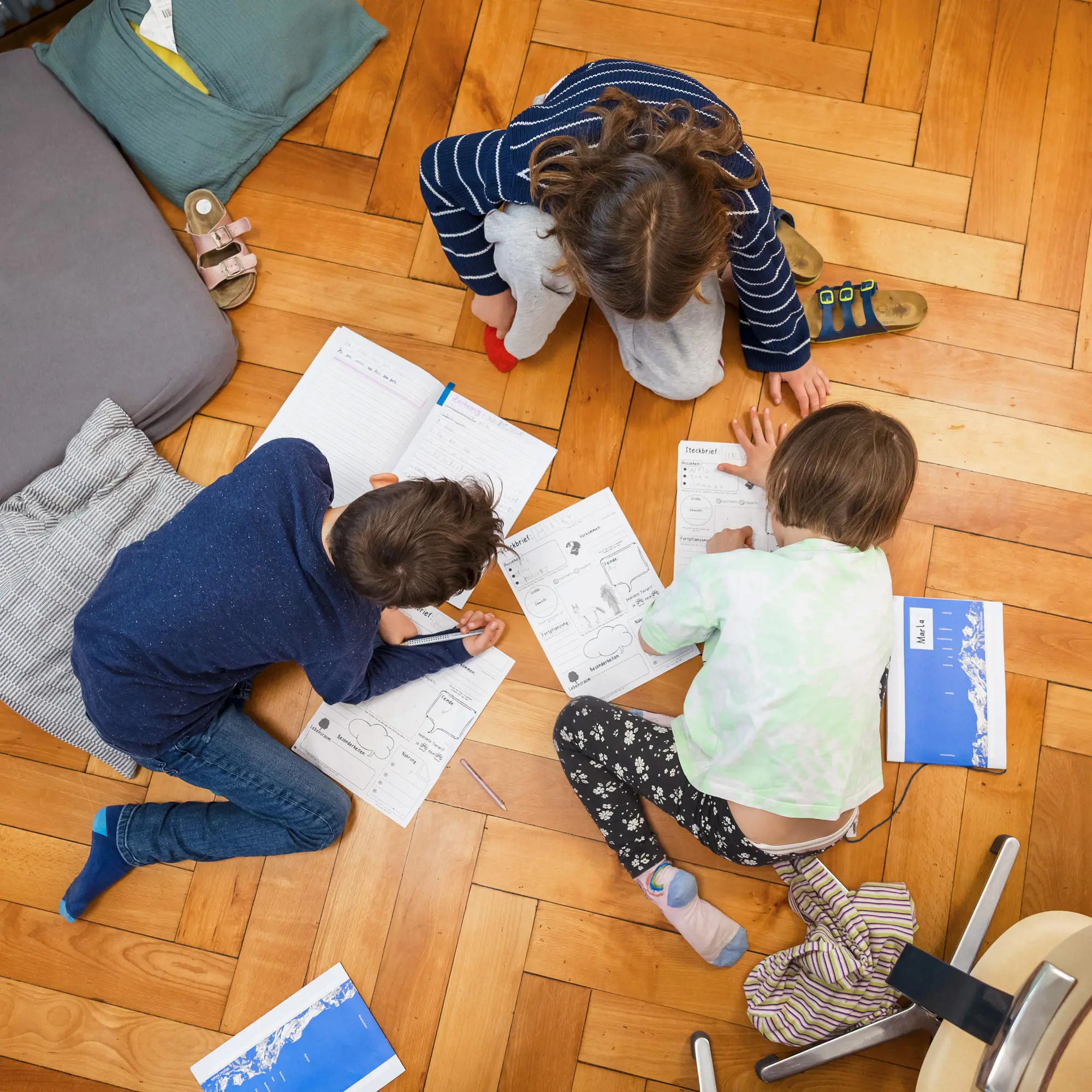 Drei Kinder arbeiten aus der Vogelperspektive gesehen gemeinsam auf dem Boden an Schulunterlagen. Sie sitzen oder knien im Kreis um mehrere geöffnete Hefte und Arbeitsblätter, die wie Steckbriefe gestaltet sind, und schreiben konzentriert darauf. Um sie herum liegen Kissen, abgelegte Schuhe und Kleidungsstücke verteilt auf dem Boden.