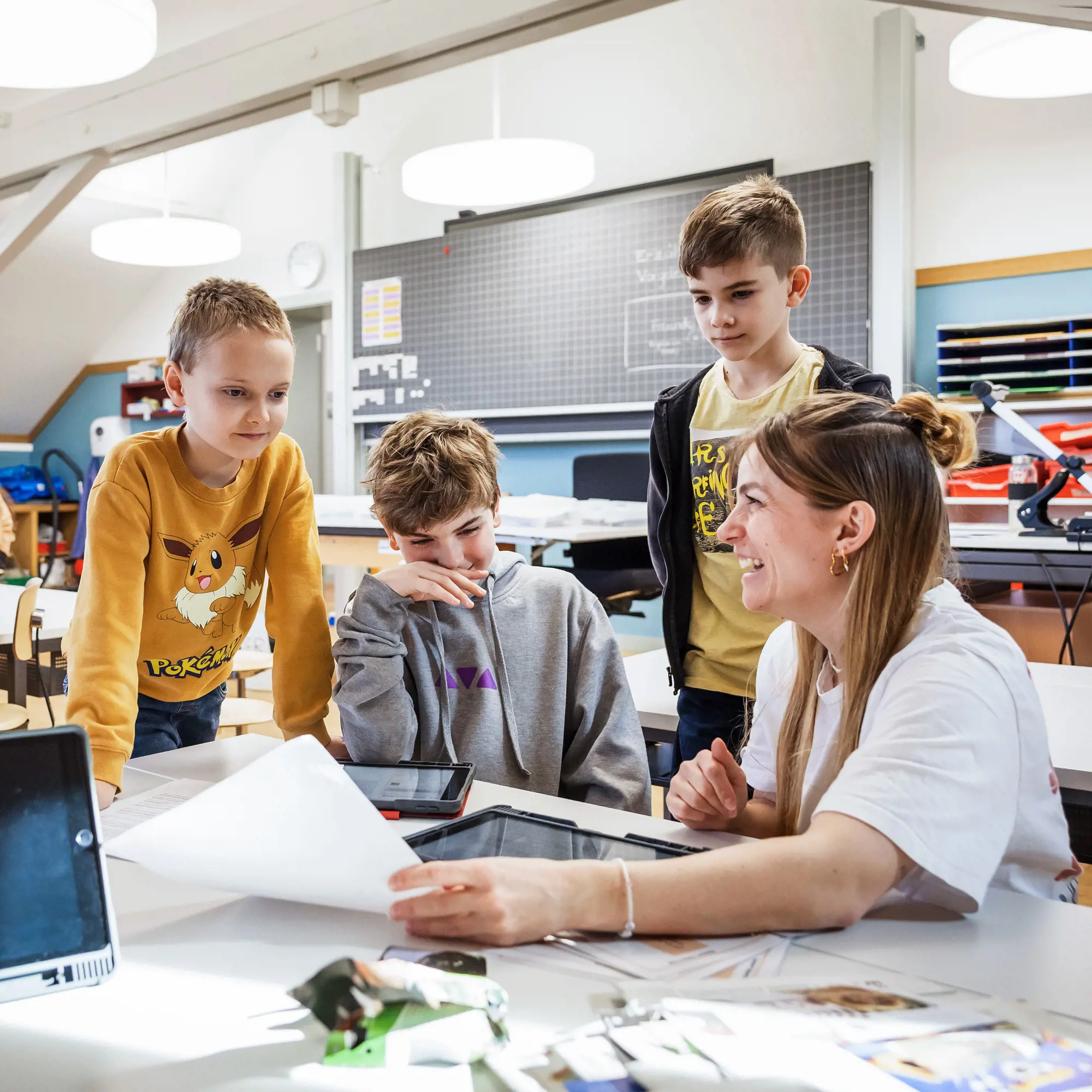 Eine Lehrerin sitzt lachend an einem Tisch im Klassenzimmer und bespricht Dokumente mit drei Jungen, die um sie herumstehen oder -sitzen. Die Gruppe blickt gemeinsam auf ein Papierblatt und auf dem Tisch liegende Tablets. Die Atmosphäre wirkt fröhlich und ungezwungen, während im Hintergrund eine Schultafel, Schränke und helle Deckenleuchten zu sehen sind.